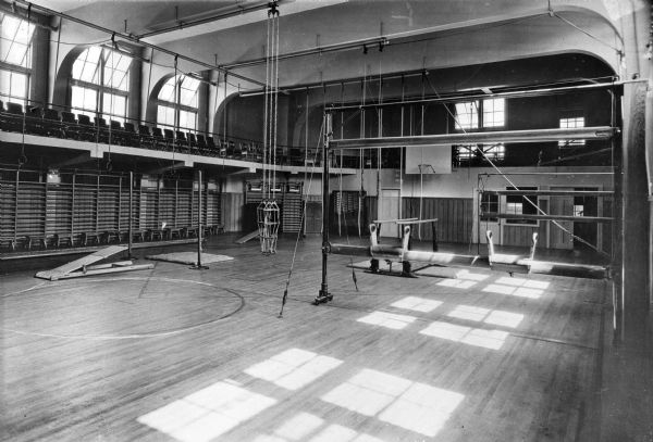 View of Y.W.C.A. Gymnasium interior equipped with climbing ropes, pummel horses, springboards, and parallel bars.  The room has a balcony with chairs for spectators.