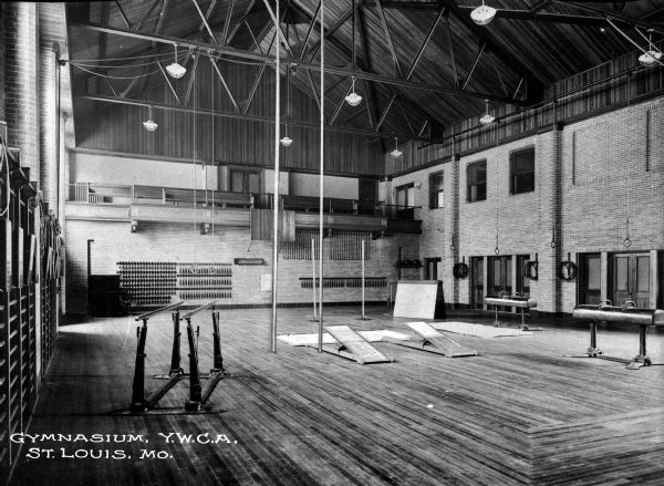 View of Y.W.C.A. gymnasium interior. View features athletic equipment including rings, springboards, and parallel bars. View also features a balcony overhead and wood floor.