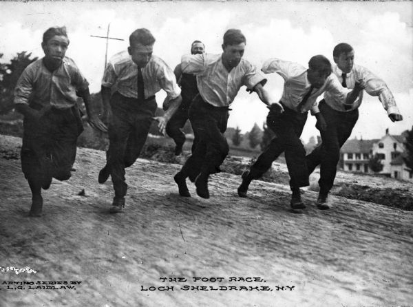 Five men compete in a foot race along a dirt road.  Buildings are visible in the background.