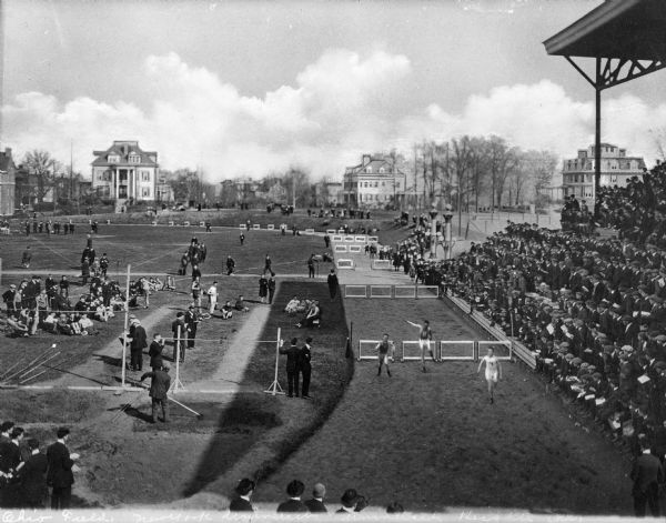 A New York University hurdle race takes place on the track at Ohio Field.  Spectators watch from a grandstand to the right and buildings are visible in the background.