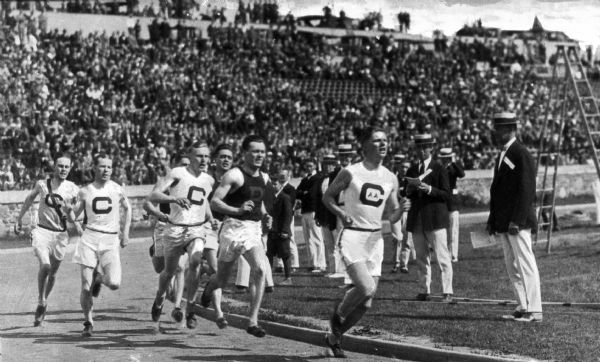 Young men in track uniforms compete in a race around a track at Cornell University.  Judges and spectators stand on the grass near the track while additional spectators watch from a grandstand in the background.