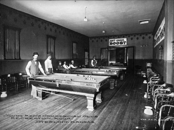 Several young men dressed in formal attire sit or play billiards at the Young Men's Commercial Club Recreation Room. The club room features three billiard tables, multiple chairs and spittoons, hardwood flooring, and a doorway with a sign above it that says "DON'T KNOCK, BOOST."