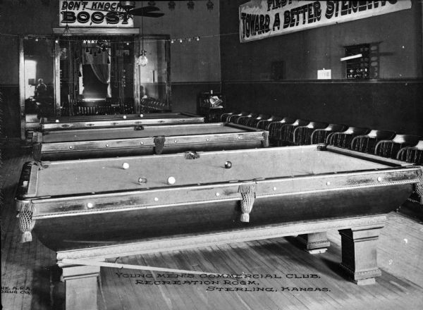 The Young Men's Commercial Club Recreation Room features three pool tables, multiple wooden chairs, several signs, hardwood floors, and an elaborate doorway.