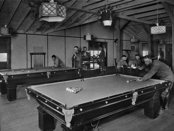 Uniformed men play billiards and relax in the Central Park Officers House. The room includes two billiard tables, chandeliers, and other interior decorations.