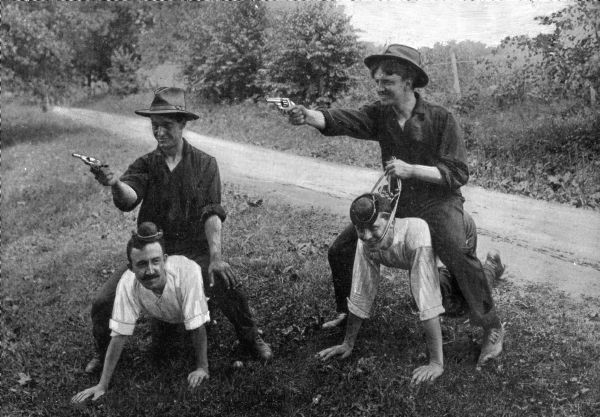 Two men crawl on their hands and knees while two other men sit atop them holding pistols. A dirt road, a fence, and foliage are in the background.