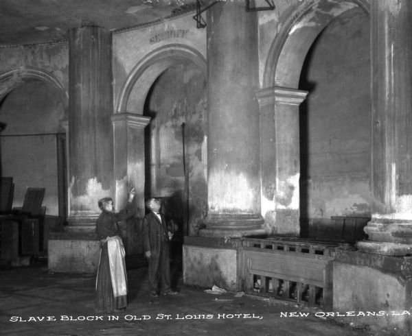 A man and a woman stand in front of archways at the Old St. Louis Hotel Slave Block. The woman points to something overhead.