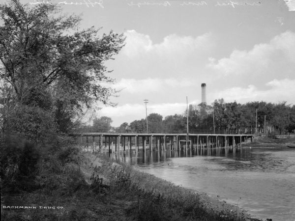 A wood bridge spans Crawfish River.  The shores are lined with foliage, power lines, and several buildings.