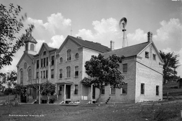 View of St. Calletta School.  The three-story brick school building features arch and rectangular windows, several porch-like entrances, and a cupola.  A windmill is visible behind the school building.