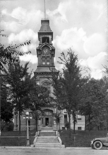An automobile parks along a road running past a courthouse.  The building features stairs leading up to the front entrance, a clock tower topped by a cupola, and rectangular windows.