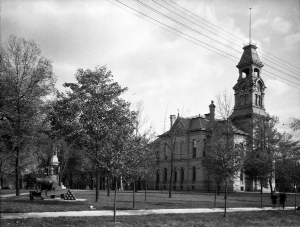A cannon and statue on a lawn next to a two-story brick building. The building features a large clock tower topped by a cupola, rectangular windows, and decorative stonework. A sidewalk runs in front of the monument and trees surround the monument and the building.