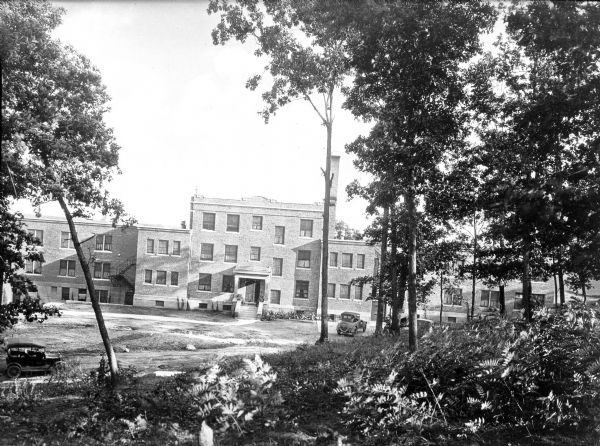 Front view of Forest Lawn Sanatorium taken from across the lawn. The brick building features rectangular windows and an entrance with stairs leading up to it. A road and several parked automobiles are in front of the building, and trees and foliage are in the foreground.