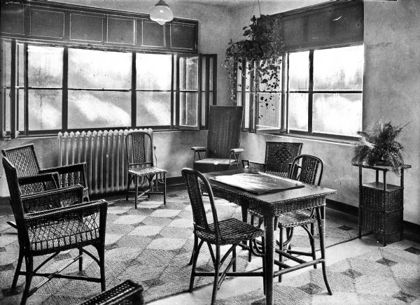 A room in Forest Lawn Sanatorium is lined on either side with windows and holds a table, end table, and several wicker chairs.  Potted plants decorate the space.