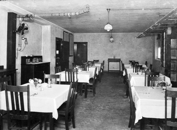 Tables set with tablecloths, floral arrangements, and silverware stand in rows inside the dining room at Forest Sanatorium dining room.
