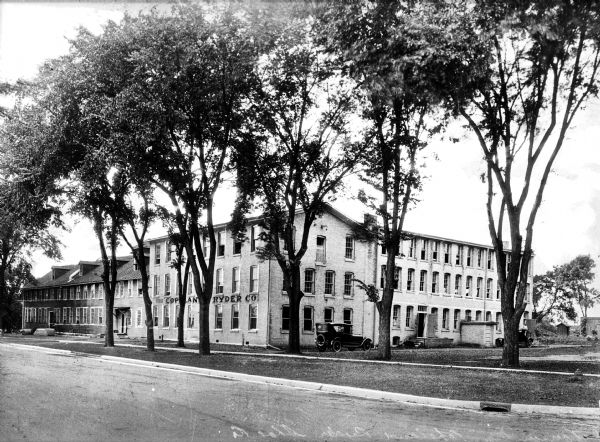 View of the exterior of the Copeland Ryder Shoe Company, established in 1868. The three-story brick building is bordered by trees, a sidewalk, and a road and two automobiles are parked along the building.