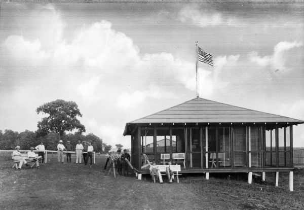 Several men stand and sit beside a building at Meadow Springs Golf Club.  The building features an elevated porch and an American flag flies from the roof.