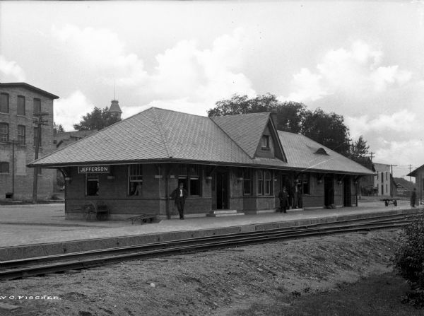 Men stand on the platform of a railroad depot while others look out from the building's windows.  Several commercial buildings stand in the background and railroad tracks pass through the foreground..