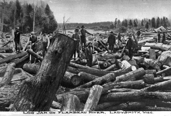 Men holding axes and other tools stand atop a log jam on the Flambeau River.