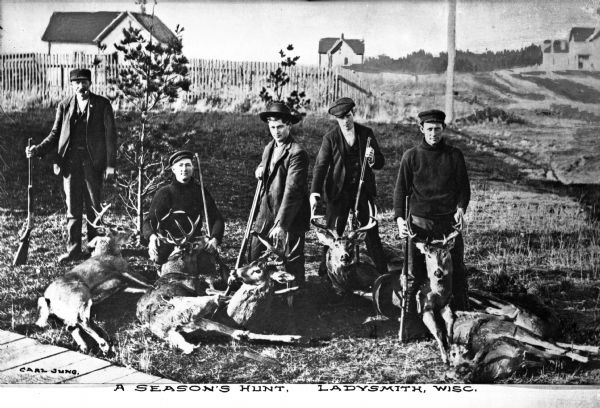 Hunters stand outdoors with deer at their feet, showing off their hunting prizes.  Several dwellings are visible in the background.