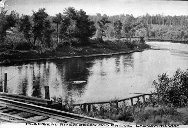 View of the Flambeau River from Soo Bridge. The railroad bridges crosses over the river and foliage lines the riverbanks.
