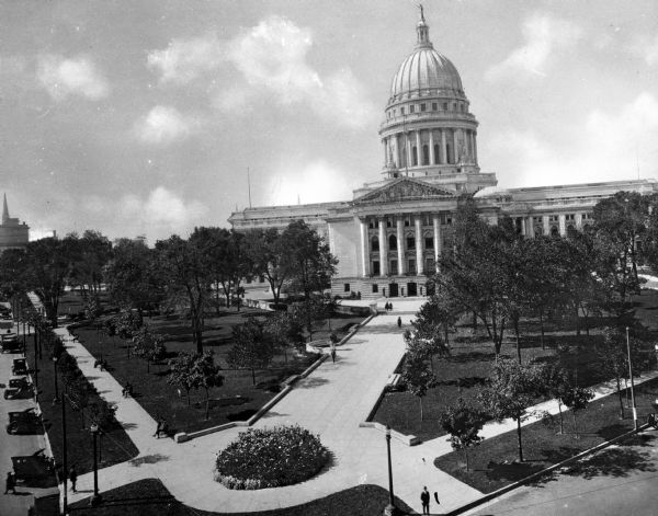Elevated view of the State Capitol looking west in Madison, Wisconsin. A corner of the Capitol Square and the Capitol Lawn are in the foreground.  Automobiles park alongside the curb and pedestrians walk along the sidewalks.