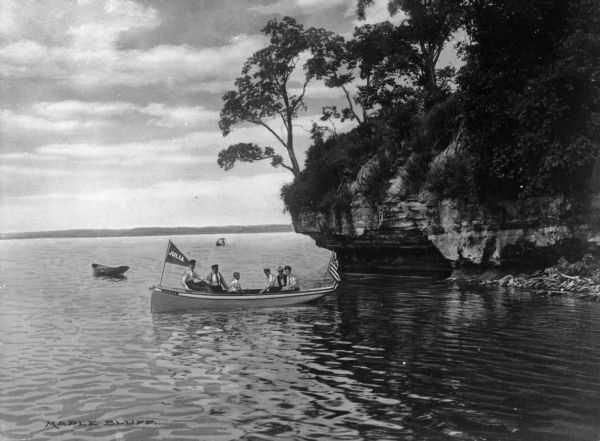 Several men and boys sail in a canoe near a foliage covered rock outcropping. The boat is decorated with two flags, an American flag and one reading "JULIA."