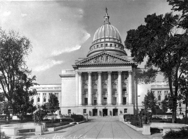 A walkway surrounded by a lawn and trees leads to the state Capitol building. The building features a large dome with Wisconsin on top, columns, a detailed pediment, arch windows, and stairs leading up to an entrance.