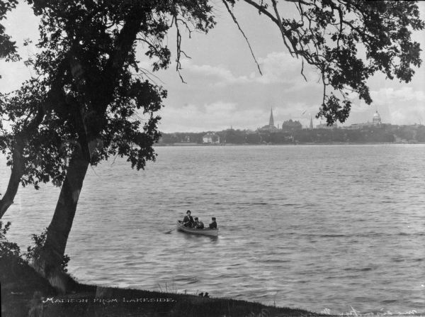 Two men and a boy row a row boat on Lake Monona.  A shoreline and trees stand in the foreground and buildings of the city of Madison can be seen in the background, including the dome of Wisconsin State Capitol building.