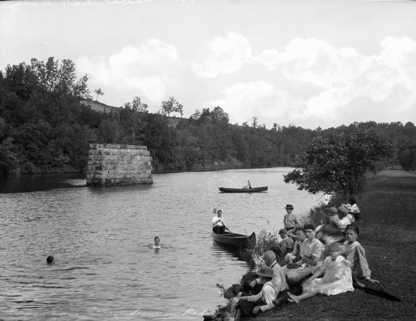 A group of youth sits on a shoreline along the Red Cedar River while others swim and boat.  A stone structure stands in the center of the river and forested shorelines surround the water.