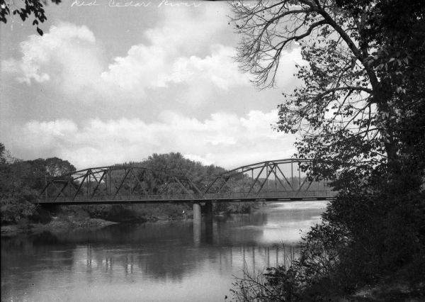 View of an arch bridge over the Red Cedar River.  Forested shorelines surround the water.