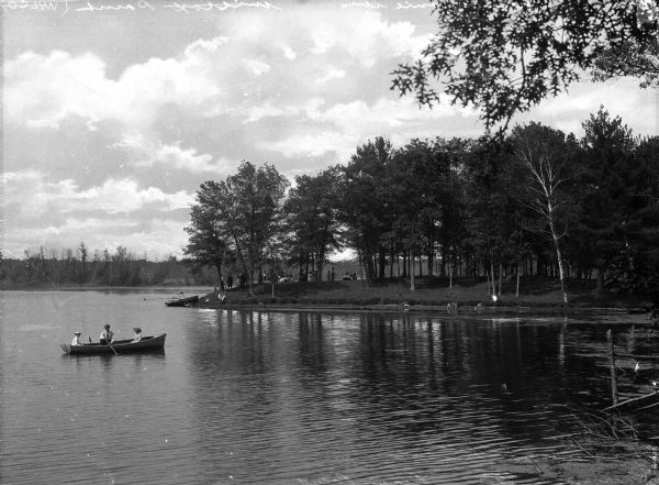 People gather on the bank of Picnic Point, a tree-covered peninsula surrounded by water.  A man and two children paddle a canoe along the shoreline.