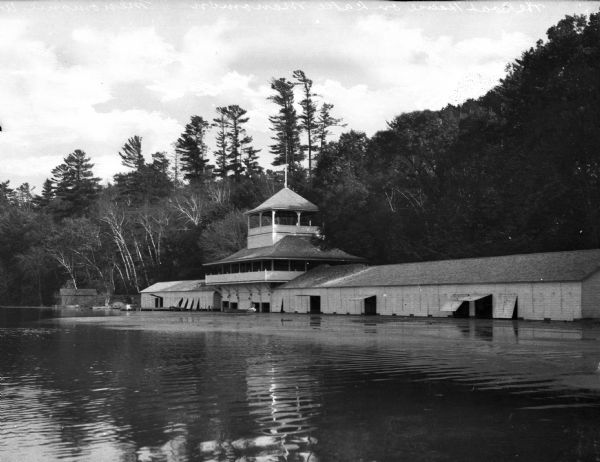 A boathouse stands along the forested shore of Lake Menomonie.  The building features multiple boat entrances and an upper-level pavilion with a lookout at center.