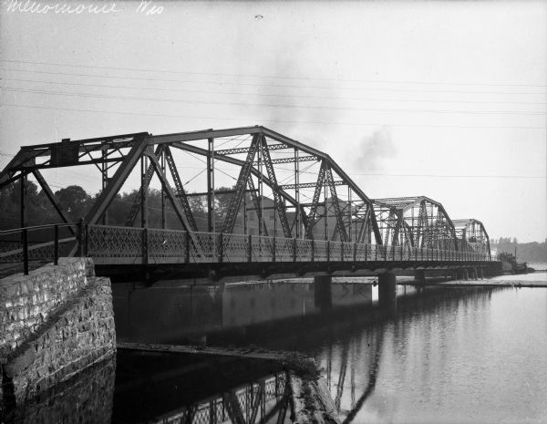 A river passes beneath a truss bridge.  City buildings are visible on the opposite side of the structure.