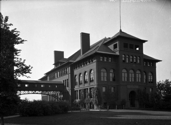 View of Central School. The brick building features arch windows and  two chimneys.  An enclosed bridge connects the building to another beside it and trees and shrubbery surround them.