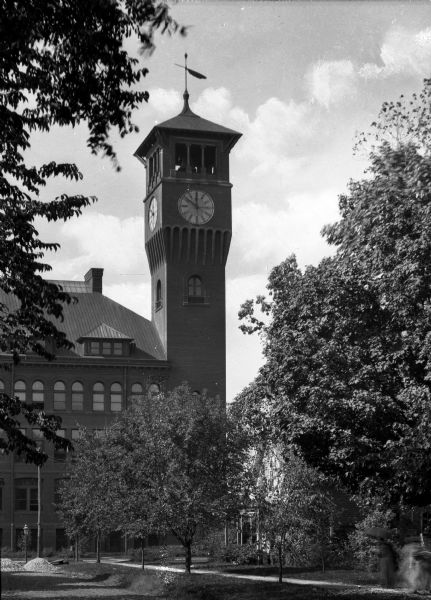 A building with an attached clock tower stands on the campus of the Stout Institute (now the University of Wisconsin-Stout).  Trees and shrubbery stand on the lawn near the building.