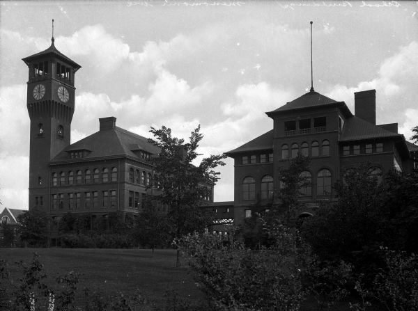 Exterior view of the manual training buildings at the Stout Institute (now the University of Wisconsin-Stout) seen from across the campus lawn. The two brick buildings are connected by a covered bridge and the building at left has a clock tower.