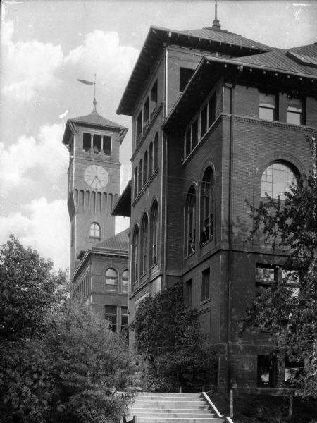 View of two brick buildings on the Stout Institute (now the University of Wisconsin-Stout) campus.  The building in the background features a clock tower and students are visible looking out of the second-floor windows of the foreground building.