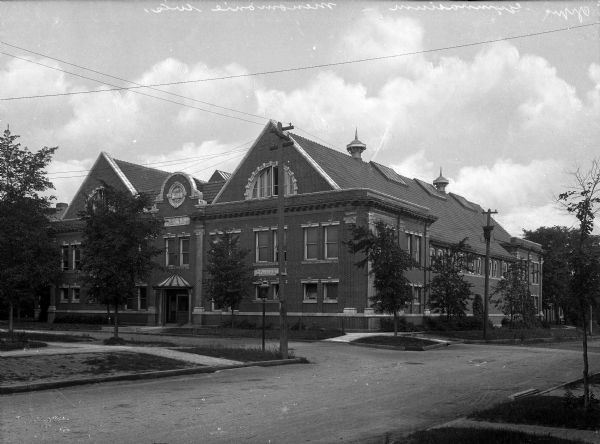 View of the Gymnasium at the University of Wisconsin-Stout, a brick building bordered by an intersection.  Several men stand at the building's entrance.