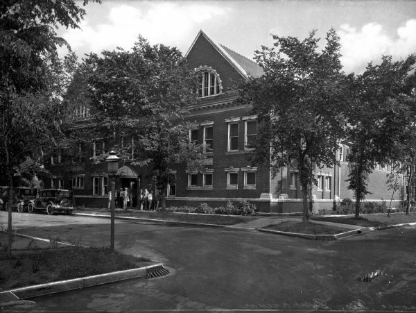 A group of men stand on a sidewalk in front of the entrance to the multi-story brick gymnasium building at the University of Wisconsin-Stout. Two automobiles park along the side of the street in front of the building and a lamppost and several shrubs and trees stand nearby.