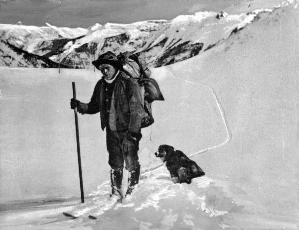 View of a mail carrier using skis and a walking stick to navigate the snow of Mount Tab. The man carries a backpack and a dog sits at his feet.