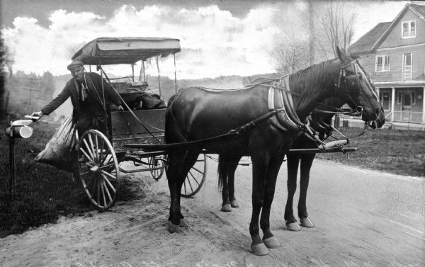 A mailman in a horse-drawn vehicle delivers mail to the mailbox of a rural boarding house.