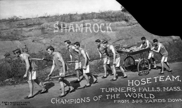 Young men on a championship hose team wear uniforms while pulling a cart along a road.