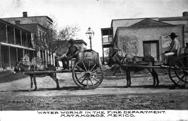 Men steer mule-drawn Fire Department carts down a dirt road in Matamoros, Mexico. Several buildings stand in the background.