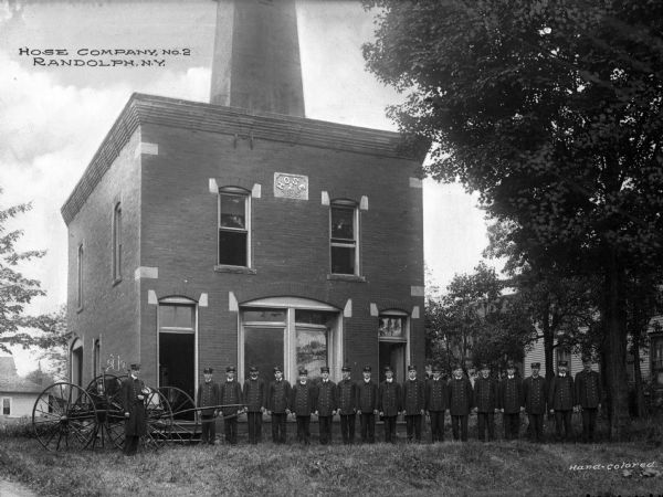 Uniformed men of Hose Company #2 fire station line up with a wagon and fire extinguishing equipment in front of the brick building.