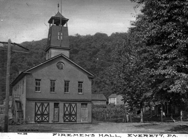 Front view of the multi-story brick Fireman's Hall.  A tower with a lookout at the top extends from the roof.  A forested hill and dwellings appear in the background.