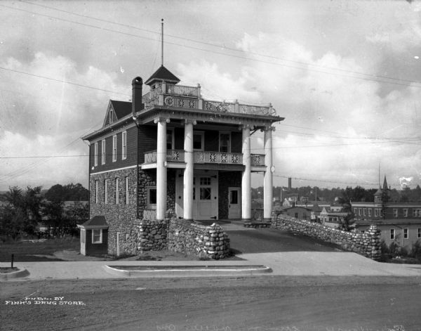 Exterior view of a firehouse building featuring two-story columns, a balcony, and stonework. A paved road leads to the structure and town buildings are visible in the background.