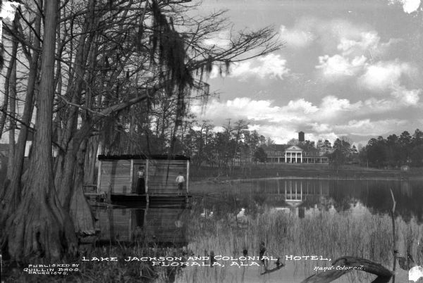 View of the Colonial Hotel as seen from across Lake Jackson. In the foreground, a woman and a boy stand next to a wooden dwelling over the water, bordered by trees. In the distance is the hotel building which features columns, a pediment, porch, and water tower. Caption reads: "Lake Jackson and Colonial Hotel, Florala, Ala."