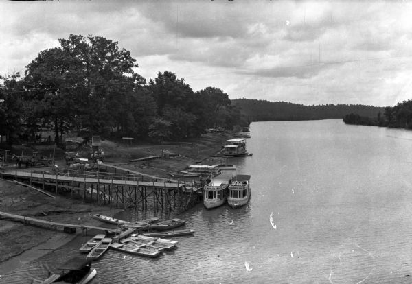 Elevated view of the Lake Taney Como boat landing featuring several piers surrounded by docked boats.  Several people stand near horse-drawn vehicles along the shoreline while others appear to be boarding a boat on the end of the largest pier.