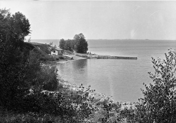 View of the lakeshore and landing at Forest View Lodge from a hillside. A small dwelling and several boats stand along the forested waterfront.