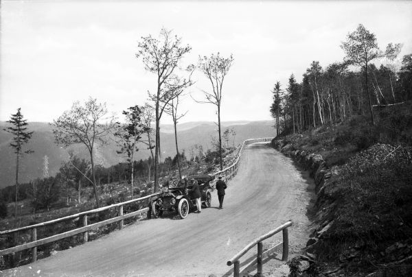 Men stand near an automobile parked along Mohawk Trail, a historic road surrounded by trees, to view a scenic overlook.