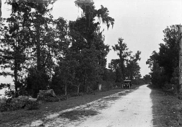 An automobile stops along Bay Shell Road, a dirt road bordered by trees and foliage on either side.